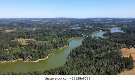 An aerial view of a serene lake surrounded by lush green forests. The calm water reflects the blue sky, and a small bridge connects two parts of the shore. - Powered by Shutterstock