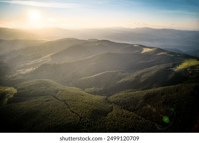 Aerial view of serene mountain landscape at sunset. Forested hills bathed in warm, golden light, creating tranquil atmosphere. Rolling hills stretch into the distance, covered in dense, lush greenery. - Powered by Shutterstock