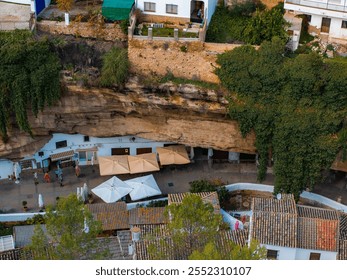 Aerial view of Setenil de las Bodegas, Spain, showcasing whitewashed buildings built under rock formations, tiled rooftops, and lush greenery. - Powered by Shutterstock