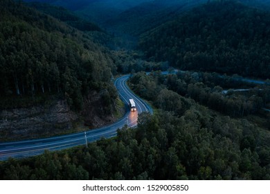 Aerial view of a sharp turn on a mountain road among green forest trees. Semi truck with cargo trailer and bright headlights on a dark highway. P-258 road near Baikal shore in Siberia, Russia