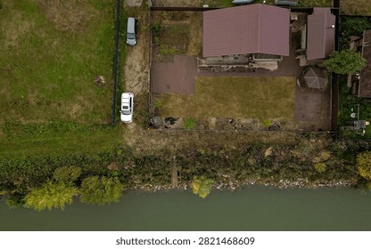 aerial view shows private plot with house featuring reddish‑brown tiled roof, paved driveway, two parked cars, small gazebo, waterfront bordered by greenery