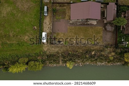 aerial view shows private plot with house featuring reddish‑brown tiled roof, paved driveway, two parked cars, small gazebo, waterfront bordered by greenery