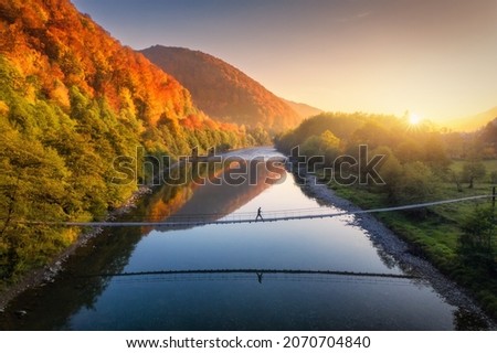 Aerial view of silhouette of walking man on the suspension bridge, river, mountains, red trees, green grass, orange sun at sunset in autumn. Colorful landscape with forest, lake, reflection in water Foto stock © 
