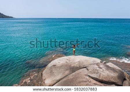 Aerial view: A slender woman stands in a bright green swimsuit with a hat in her hand stands on a rock and admires the azure sea.