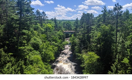 Aerial View Of A Small Rushing River With A Picturesque Bridge In The Background Surrounded By A Lush Pine Tree Forest In The Upper Peninsula Of Michigan