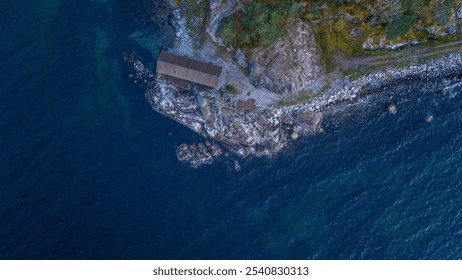 Aerial view of a small wooden cabin on a rocky coastline surrounded by deep blue ocean water and lush greenery. - Powered by Shutterstock