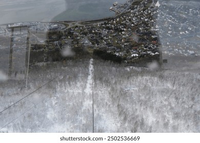 Aerial view of a snow-covered hill descending into a coastal town by the water, with the town's structures and streets visible amidst the winter landscape. - Powered by Shutterstock