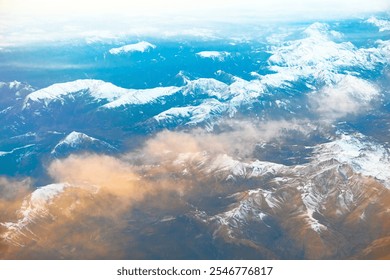 Aerial view of snow-covered mountain peaks partially obscured by clouds during sunrise. Rugged terrain contrasts with the soft clouds highlighting the natural beauty and grandeur of the landscape. - Powered by Shutterstock