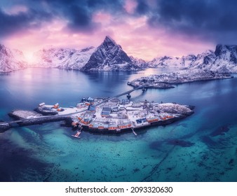 Aerial view of snowy islands with houses, rorbu, blue sea, mountains, bridge and violet cloudy sky at sunset in winter. Dramatic landscape with village, rock, road. Top view. Lofoten islands, Norway