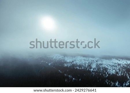 Aerial view of snowy mountain range in cloud with sun shining through during frosty day. Moody winter scenery.
