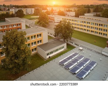 Aerial View Of Solar Power Plant Instaled On The School Building Roof Against Cityscape And Orange Sunset.