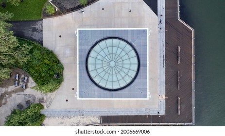 An aerial view of a square structure with a round, glass rooftop that looks like a spiderweb. Taken by the East River it has a concrete foundation, surrounded by a beautiful landscape on a sunny day.