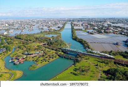 Aerial View Of A Steam Locomotive Hauling A Train Thru A Bridge Over A Forest Park By Dongshan River With Houses Scattered Amid Irrigated Paddies And Turtle Island On Horizon In Luodong, Yilan, Taiwan