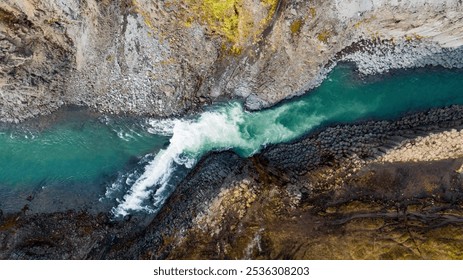 Aerial view of a steep, narrow Icelandic canyon with turquoise water rushing through it, flanked by rugged, rocky slopes and a grassy plateau under a cloudy sky, showcasing untouched wilderness. - Powered by Shutterstock