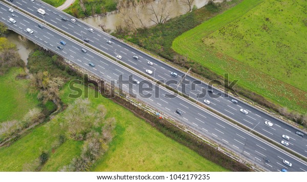 Aerial view of a stretch of highway in the Italian\
countryside. The road is divided into many lanes in each direction.\
There are cars and trucks on the street.Around the road there are\
trees and grass