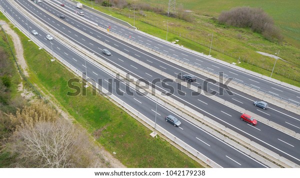 Aerial view of a stretch of highway in the Italian\
countryside. The road is divided into many lanes in each direction.\
There are cars and trucks on the street.Around the road there are\
trees and grass