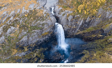 Aerial view of a stunning waterfall cascading down rocky cliffs into a deep blue pool surrounded by lush greenery and autumn foliage. - Powered by Shutterstock