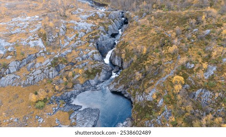 Aerial view of a stunning waterfall cascading down rocky cliffs into a deep blue pool surrounded by lush greenery and autumn foliage. - Powered by Shutterstock