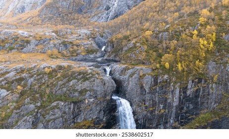 Aerial view of a stunning waterfall cascading down rocky cliffs into a deep blue pool surrounded by lush greenery and autumn foliage. - Powered by Shutterstock