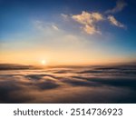 Aerial view of sun rises above sea of clouds, painting sky with soft hues of orange and blue. Light, wispy clouds scatter across horizon, capturing serene and beauty of dawn breaking over landscape.