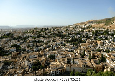 Aerial view of a sunlit Mediterranean city with tightly packed historic buildings, whitewashed facades, and lush greenery, set against rolling hills. - Powered by Shutterstock