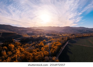 An aerial view of a sunset over Foxen Canyon Road and Foxen Winery in Santa Ynez, California. Golden hour light bathes the rolling hills, vineyards, and autumn-colored trees in a picturesque scene. - Powered by Shutterstock