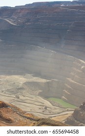 Aerial View Of Super Pit Goldmine In Kalgoorlie, Western Australia, With Winding Path Along Edges Of The Whole, Heavy Machinery And Groundwater Lake At Bottom.