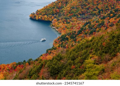 Aerial view of a tour boat cruising on magnificent Lake Chuzenji (中禅寺湖, a famous tourist destination in Nikko National Park, Tochigi Prefecture, Japan), with brilliant fall colors on lakeside hills