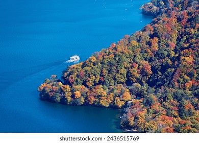 Aerial view of a tour boat cruising on Lake Chuzenji 中禅寺湖 on a sunny autumn day, in Nikko National Park, Tochigi Prefecture, Japan, with beautiful fall colors on the lakeside hills and Hachodeshima
