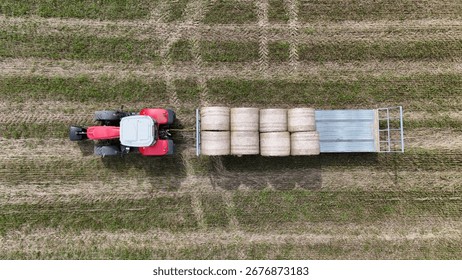 Aerial view of tractor pulling trailer loaded with hay bales across farmland, representing rural work, agriculture, and harvest efficiency.