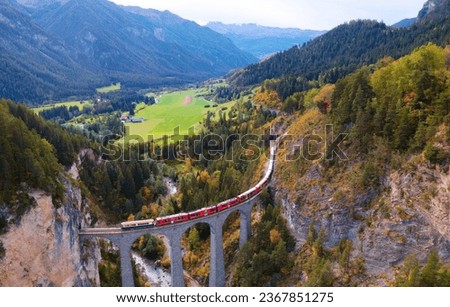 Aerial view of a train of Rhaetian Railway (RhB) winding thru fall colors and crossing Landwasser Viaduct over a gorge to enter a tunnel in the cliff, in Filisur, Grisons (Graubünden), Switzerland