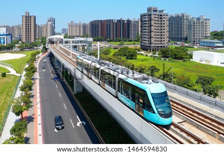 Aerial view of a train traveling on the elevated track of Danhai Light Rail System near Tamsui District Office Station with residential towers standing under sunny sky in background in New Taipei City