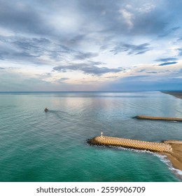 Aerial view of a tranquil coastal harbor with breakwaters and a small boat.A serene aerial shot of a coastal harbor with calm turquoise water, a small boat sailing, and two breakwaters extending into  - Powered by Shutterstock