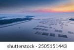 Aerial view of tranquil coastal waters at sunset, dotted with geometric seaweed farms, distant wind turbines on land, creating harmonious contrast.