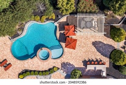 Aerial View Of Trendy Curving Pool With Turquoise Water, White Outdoor Patio Pergola Shade Structure With Seating, Dining Table, Orange Beach Chairs, Curved Landscaping, Orange Beach Umbrellas