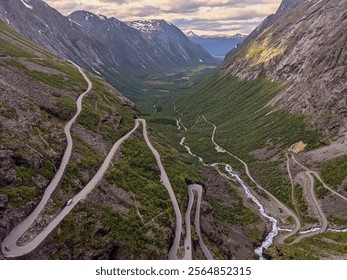 Aerial view of Trollstigen, Norway, featuring winding mountain roads, steep cliffs, lush green valleys, and cascading waterfalls under a cloudy sky. A dramatic scenic route through rugged terrain - Powered by Shutterstock