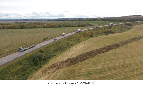 Aerial View Of Trucks On Road Driving Away In Beautiful Countryside With Green Grass In Direction Of Loading Warehouse Area. Large Delivery Truck Is Moving.