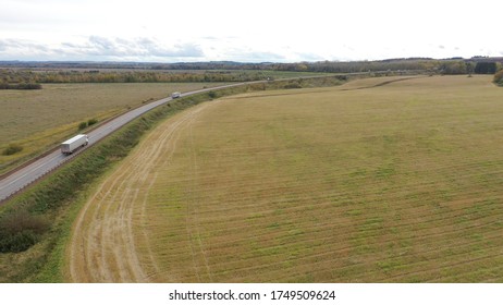 Aerial View Of Trucks On Road Driving Away In Beautiful Countryside With Green Grass In Direction Of Loading Warehouse Area. Large Delivery Truck Is Moving.