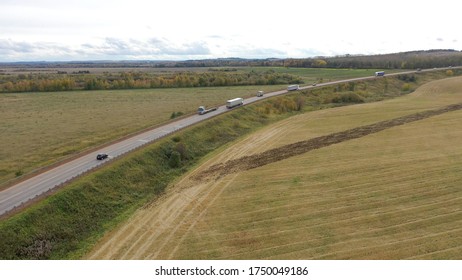 Aerial View Of Trucks On Road Driving Away In Beautiful Countryside With Green Grass In Direction Of Loading Warehouse Area. Large Delivery Truck Is Moving.