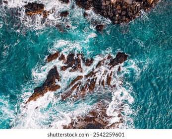 Aerial view of Turquoise waves crash against rocky coastline during sunny daylight - Powered by Shutterstock