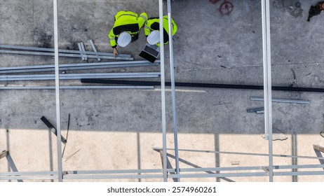 Aerial view of two construction workers in safety gear reviewing plans on a laptop amidst metal beams, emphasizing teamwork, precision, and the role of technology in modern construction projects.