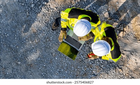 Aerial view of two construction workers in safety gear reviewing plans on a laptop amidst metal beams, emphasizing teamwork, precision, and the role of technology in modern construction projects.