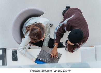 An aerial view of two professionals working together at a white desk, reviewing content on a laptop in a bright, contemporary office setting, highlighting teamwork and cooperation.