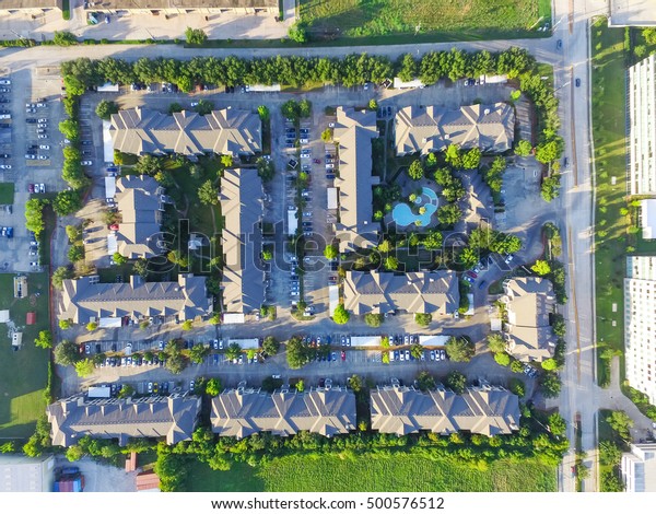 Aerial view of typical multi-level apartment\
building with swimming pool, surrounded by green garden and rows of\
cars in parking lots in Houston, Texas, US. Apartment complex next\
to road at sunset.