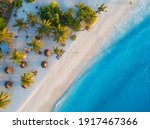 Aerial view of umbrellas, palms on the sandy beach of Indian Ocean at sunset. Summer in Zanzibar, Africa. Tropical landscape with palm trees, parasols, walking people, blue water, waves. Top view