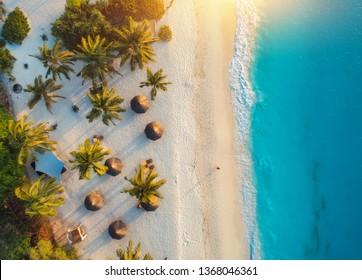 Widok z lotu ptaka parasole, palmy na piaszczystej plaży Oceanu Indyjskiego o zachodzie słońca. Letnie wakacje w Zanzibarze, Afryka. Tropikalny krajobraz z palmami, parasolami, białym piaskiem, błękitną wodą, falami. Widok z góry