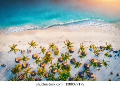 Aerial View Of Umbrellas, Palms On The Sandy Beach Of Indian Ocean At Sunset. Summer Holiday In Zanzibar, Africa. Tropical Landscape With Palm Trees, Parasols, White Sand, Blue Water, Waves. Top View