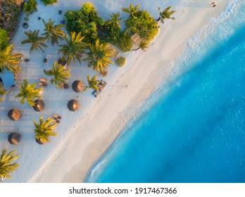 Aerial View Of Umbrellas, Palms On The Sandy Beach Of Indian Ocean At Sunset. Summer In Zanzibar, Africa. Tropical Landscape With Palm Trees, Parasols, Walking People, Blue Water, Waves. Top View