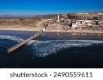 Aerial view of the US-Mexico border wall extending into the Pacific Ocean at Tijuana Beach. The scene captures the contrast between the bustling urban area and the serene beachfront.