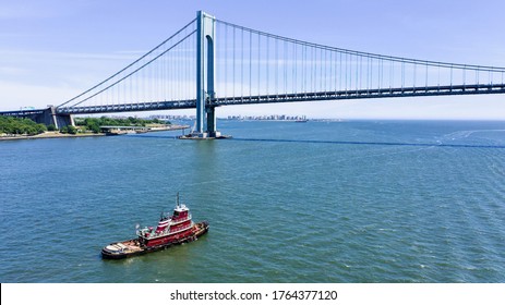 An aerial view of the Verrazano bridge, with a tug boat in the forefront of the frame. The drone is hovering over New York Bay to capture the shot. It is a bright and sunny day with clear blue skies.